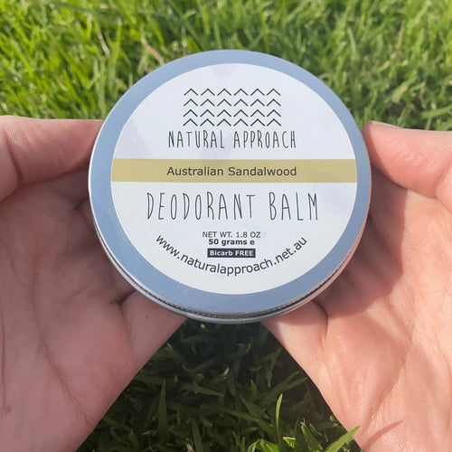 Hand holding a container of Natural Approach Australian Sandalwood Deodorant Balm against a grass background