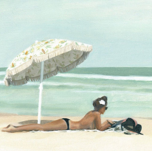 The image shows a woman laying on the beach, raised up on her elbows. She is enjoying the sunshine under an umbrella, and appears to be reading a book or looking into the distance.