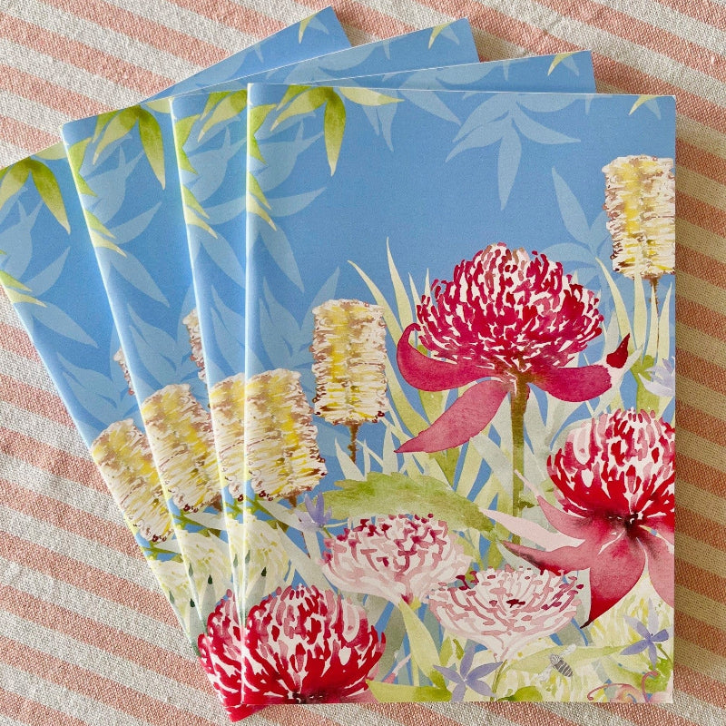 The image shows the illustrated artwork on the A5 Notebook by Fabriculture. The Artist is Penelope Gale. The Notebook has a blue background and features the bright red Gibraltar Range Waratah amongst some foliage and bottlebrush.