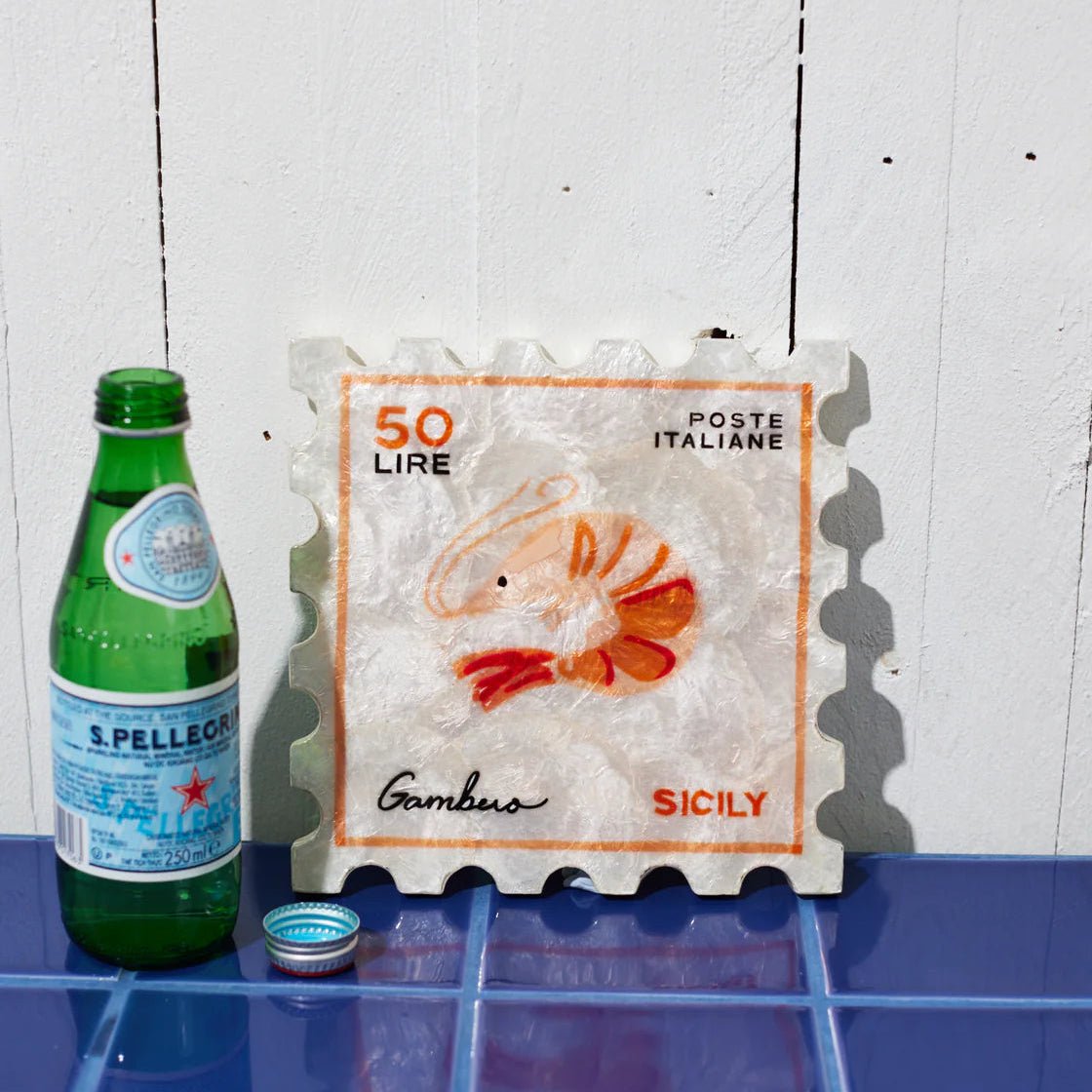 Green bottle with a label next to a postage stamp piece of wall art, of a prawn, on a tiled surface